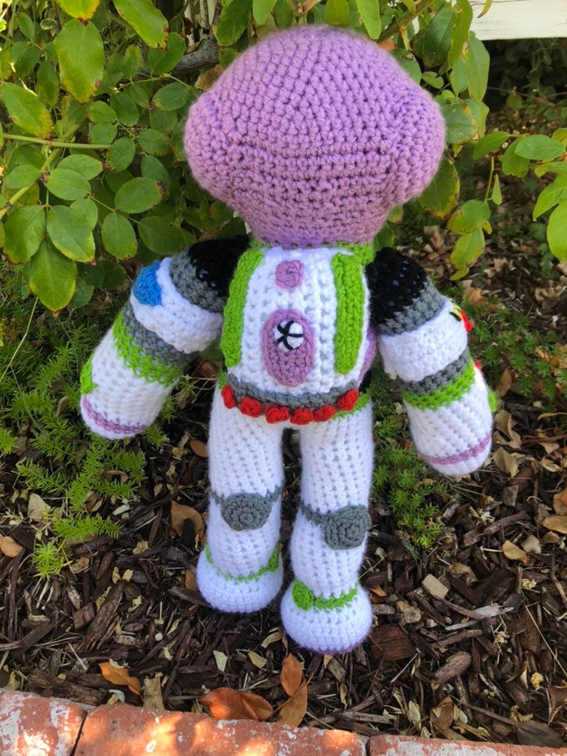 May include: A crocheted plush toy of Buzz Lightyear from the Toy Story franchise. The toy is purple, white, green, and gray. The toy is standing on a bed of mulch.