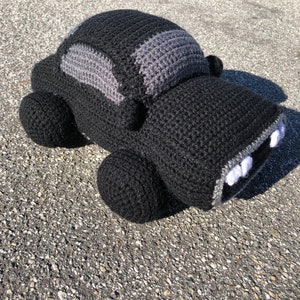 Peut inclure: Un jouet de voiture en crochet noir avec des détails gris. La voiture a des dents blanches et un toit gris.