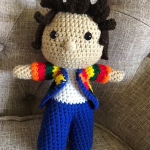 Puede incluir: Una muñeca de crochet con pelo marrón, una camisa blanca, pantalones azules y una chaqueta de rayas de colores.