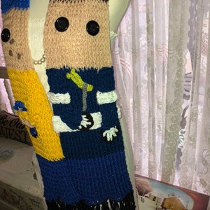 May include: A knitted scarf with a cartoon face on it. The scarf is blue, yellow, brown, and white. It has black fringe on the bottom.