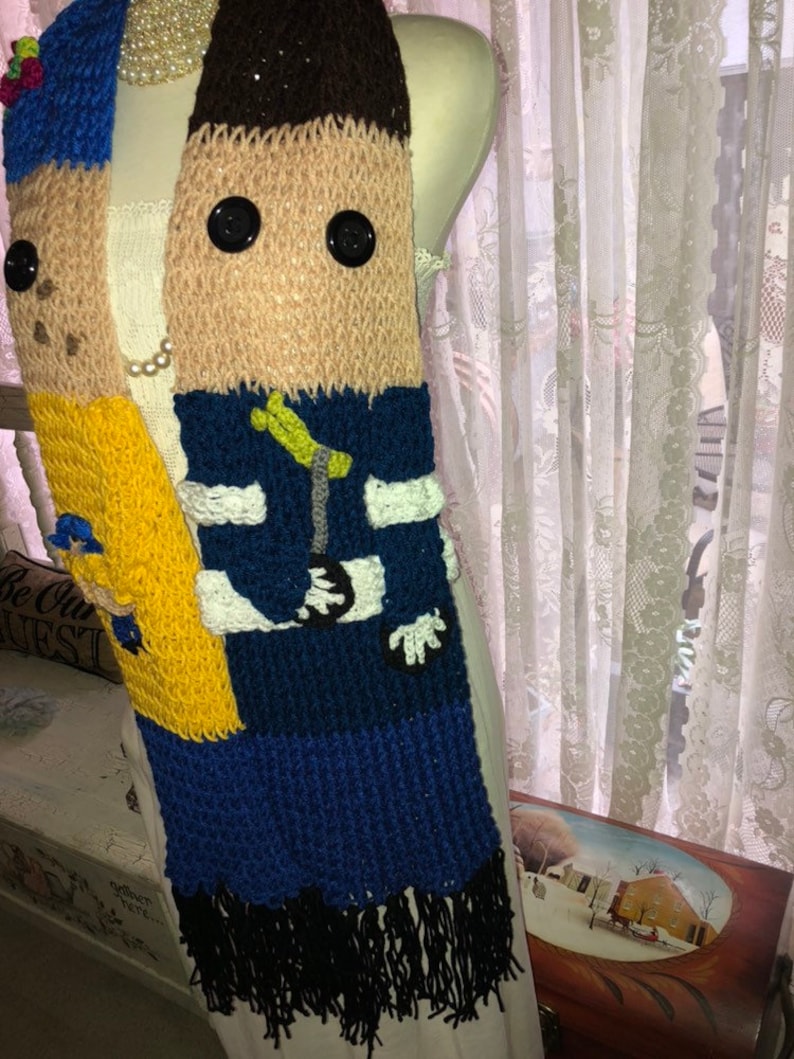 May include: A knitted scarf with a cartoon face on it. The scarf is blue, yellow, brown, and white. It has black fringe on the bottom.