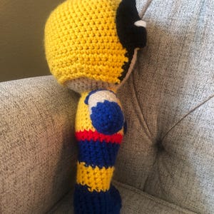 Op de afbeelding: Handgemaakte gehaakte pop van een superheld, met een gele kop en kap met zwarte accenten. Het lichaam is een combinatie van blauw, geel en rood garen, met een grijze basis. De pop zit op een grijs gestoffeerde stoel.
