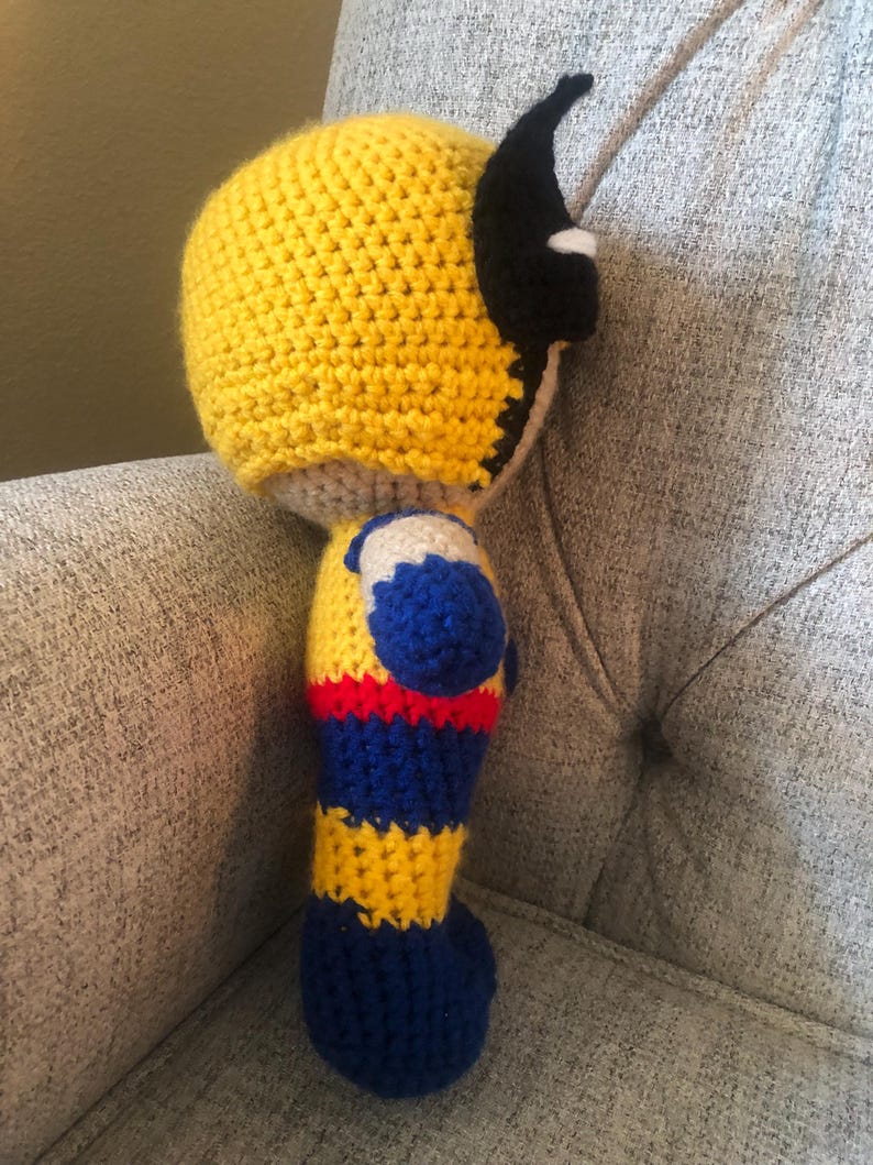 Op de afbeelding: Handgemaakte gehaakte pop van een superheld, met een gele kop en kap met zwarte accenten. Het lichaam is een combinatie van blauw, geel en rood garen, met een grijze basis. De pop zit op een grijs gestoffeerde stoel.