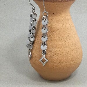 Może przedstawiać: Para srebrnych kolczyków z łańcuszkiem małych okrągłych dysków i diamentowym zawieszką na dole.