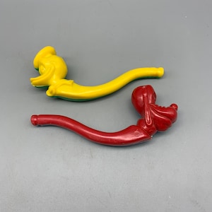 Peut inclure: Deux pipes en plastique pour enfants, l'une jaune et l'autre rouge. La pipe jaune a une tête de canard de dessin animé à l'extrémité et la pipe rouge a une tête de chien de dessin animé à l'extrémité.