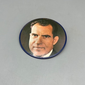 May include: A close-up of a round button featuring a colour photograph of Richard Nixon. The button has a blue border.