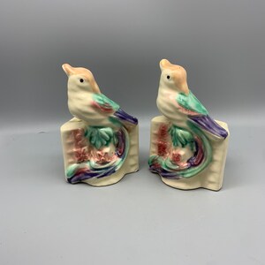 Peut inclure: Une paire de serre-livres en céramique en forme d'oiseaux avec une base blanche et des détails peints colorés. Les oiseaux sont perchés sur un motif floral stylisé avec des accents roses, violets et verts.
