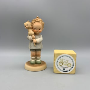 May include: A porcelain figurine of a girl holding a small kitten. The figurine is painted in soft colours and has a delicate, vintage style. The figurine is standing on a small base and is accompanied by a wooden box with a bicycle logo.