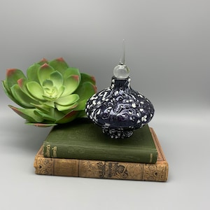 Pode incluir: Uma garrafa de vidro azul escuro e branco com um design de redemoinho e um tampão de vidro transparente. A garrafa está em cima de dois livros.