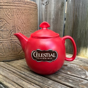 May include: A red ceramic teapot with a black logo that reads "Celestial Seasonings" on a wooden surface.