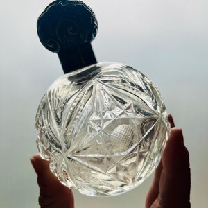 May include: A clear glass bottle with a black, ornate stopper. The bottle has a faceted, geometric design.