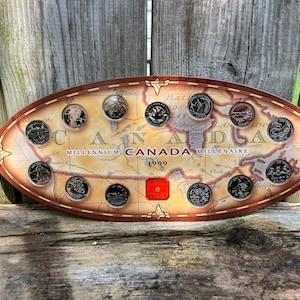 May include: A collection of 10 Canadian coins from 1999, each featuring a different province or territory, displayed in a circular frame with a map of Canada. The frame is brown with gold accents and the text "Millennium Canada Millenaire 1999".