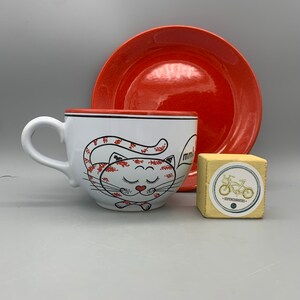 May include: A white ceramic teacup featuring a red and white cat design. The cat has its eyes closed and a red heart on its chest. The cup is accompanied by a red saucer and a small wooden cube with a bicycle sticker.