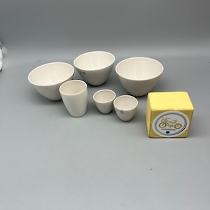 Könnte beinhalten: Ein Set aus sechs weißen Keramikschalen in verschiedenen Größen, darunter ein kleiner gelber Würfel mit einem Aufkleber eines Fahrrads.