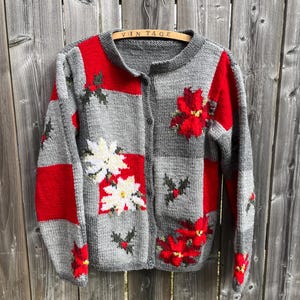 May include: A gray knitted cardigan with red and white poinsettia flowers and holly leaves. The cardigan has a button closure and is hanging on a wooden hanger labeled "Vintage".