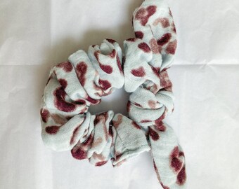 May include: A light blue scrunchie with a maroon and tan spotted pattern. The scrunchie has a knotted bow detail. The fabric is soft and appears to be cotton. This hair accessory is suitable for various hairstyles.