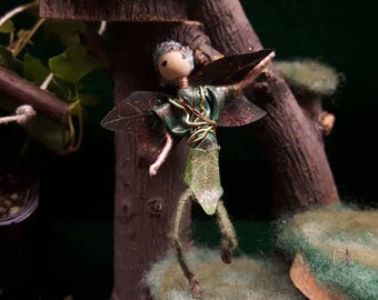 May include: A whimsical fairy figurine with green wings, a green and brown dress, and a wire frame. The fairy is posed with its arms outstretched and its head tilted to the side. The figurine is made of natural materials and has a rustic charm.