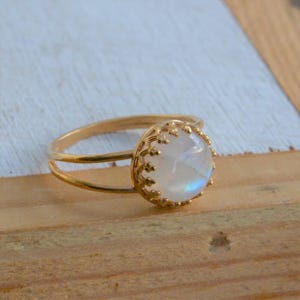 Puede incluir: Un anillo de oro con una piedra de luna redonda y blanca engastada en un delicado marco en forma de corona.