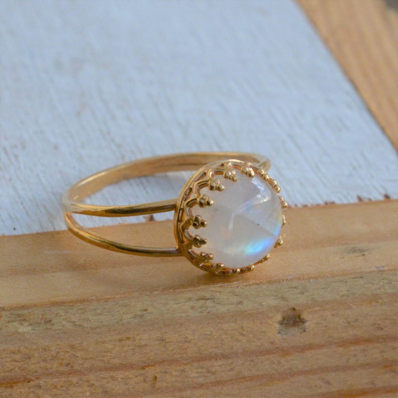 Puede incluir: Un anillo de oro con una piedra de luna redonda y blanca engastada en un delicado marco en forma de corona.