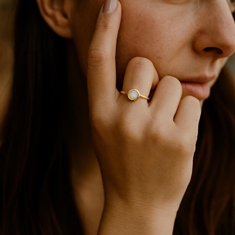 May include: A gold ring with a round, light-colored stone. The ring is on a person's finger, with the hand in focus. The ring's band is thin and simple, and the stone is set in a bezel setting. The background is blurred.