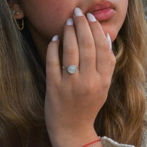 Puede incluir: Un anillo de plata con una piedra de luna blanca engastada en un bisel festoneado. El anillo est&aacute; en el dedo de una persona.