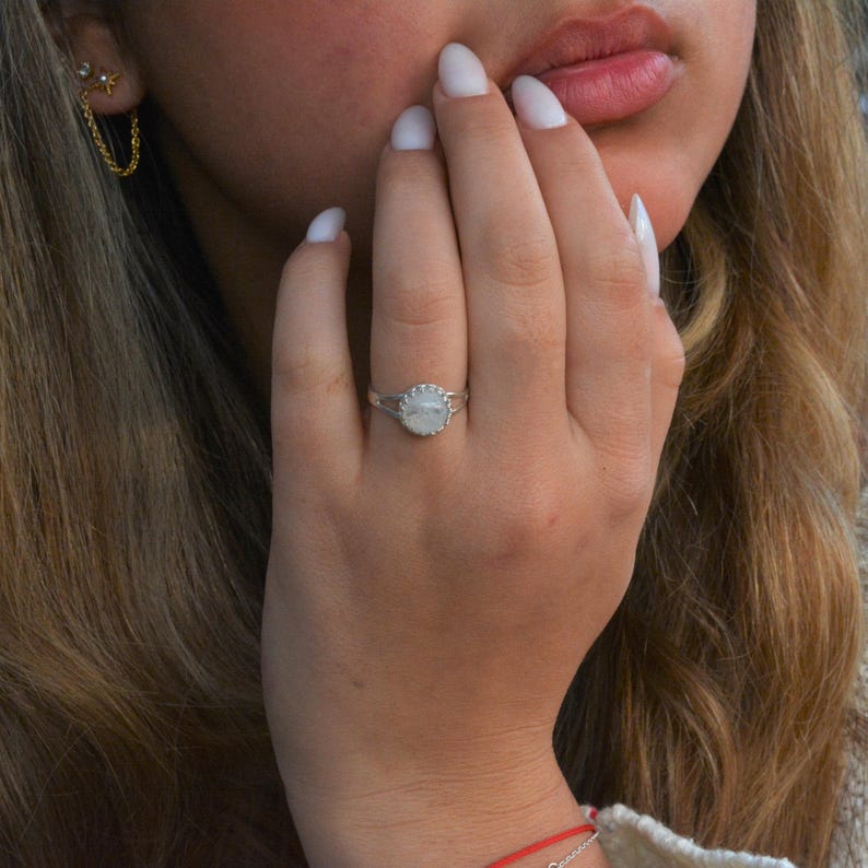 Puede incluir: Un anillo de plata con una piedra de luna blanca engastada en un bisel festoneado. El anillo est&aacute; en el dedo de una persona.
