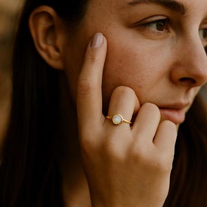 May include: A gold ring with a round, light-colored stone. The ring is on a finger, and the person is touching their face. The ring's band is thin and simple, and the stone is set in a bezel.