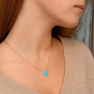 May include: A gold-colored necklace with a small, turquoise-colored hand-shaped pendant. The necklace is worn around the neck. The background is blurred.