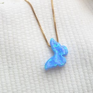 May include: A gold-colored chain necklace with a blue butterfly pendant. The pendant has a shimmering, opalescent appearance with shades of blue and green. The necklace is displayed on a white, textured surface.
