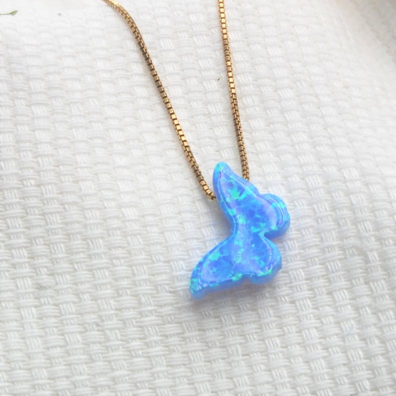 May include: A gold-colored chain necklace with a blue butterfly pendant. The pendant has a shimmering, opalescent appearance with shades of blue and green. The necklace is displayed on a white, textured surface.