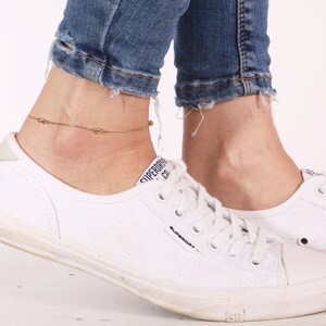 May include: A gold anklet with small round beads on a woman's ankle. The anklet is worn with white sneakers and blue jeans.