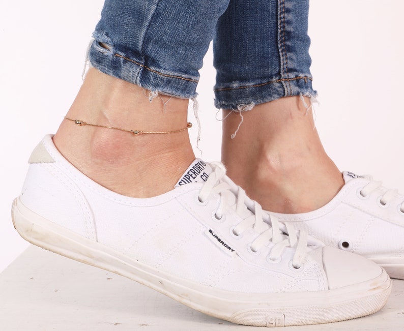 May include: A gold anklet with small round beads on a woman's ankle. The anklet is worn with white sneakers and blue jeans.