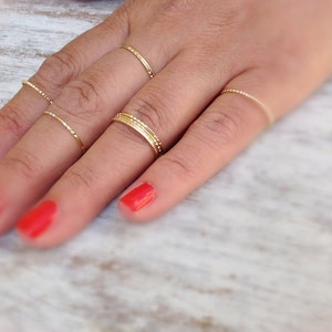 May include: A hand wearing multiple thin gold rings on different fingers. The rings are delicate and simple, with a subtle shine. The nails are painted a bright red.