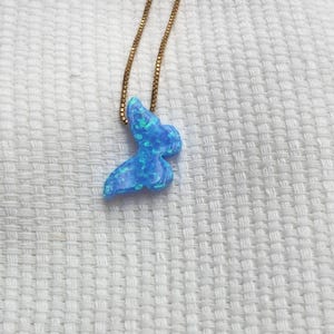 May include: A blue opal butterfly pendant necklace with a gold chain. The butterfly pendant has a shimmering, iridescent quality with shades of blue and green. The necklace is displayed on a white textured surface.