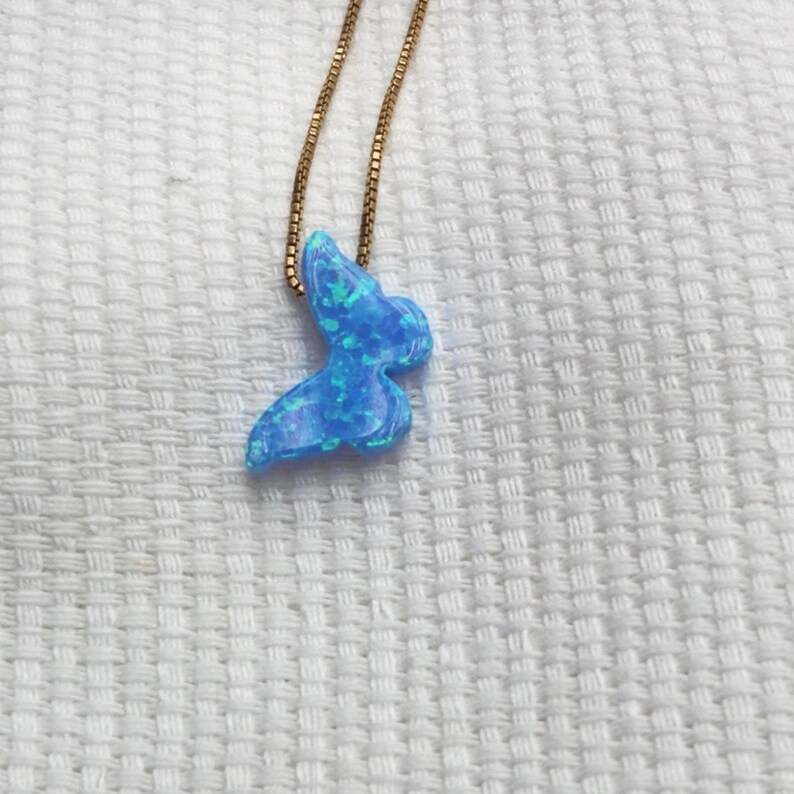 May include: A blue opal butterfly pendant necklace with a gold chain. The butterfly pendant has a shimmering, iridescent quality with shades of blue and green. The necklace is displayed on a white textured surface.