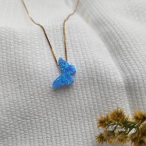 May include: A delicate gold-tone necklace featuring a blue butterfly pendant. The butterfly pendant has a shimmering, opalescent appearance. The necklace is displayed on a white, textured surface, with a sprig of yellow flowers in the background.