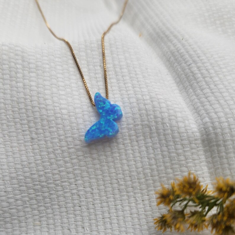 May include: A delicate gold-tone necklace featuring a blue butterfly pendant. The butterfly pendant has a shimmering, opalescent appearance. The necklace is displayed on a white, textured surface, with a sprig of yellow flowers in the background.