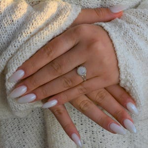 Puede incluir: Un anillo de plata con una piedra de luna blanca engastada en un dise&ntilde;o delicado y ornamentado. El anillo se lleva en una mano con u&ntilde;as largas, ovaladas y blancas.