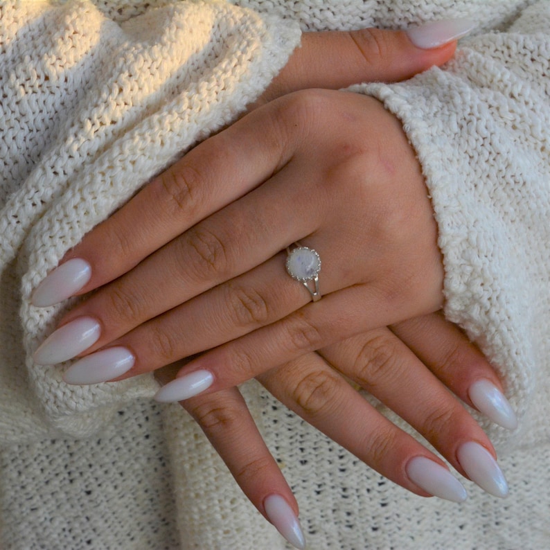 Puede incluir: Un anillo de plata con una piedra de luna blanca engastada en un dise&ntilde;o delicado y ornamentado. El anillo se lleva en una mano con u&ntilde;as largas, ovaladas y blancas.