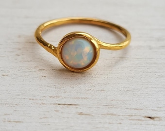 May include: A gold-colored ring featuring a round, iridescent opal stone. The opal is set in a bezel setting, and the band has a textured, hammered appearance. The ring is displayed on a light-colored wooden surface.