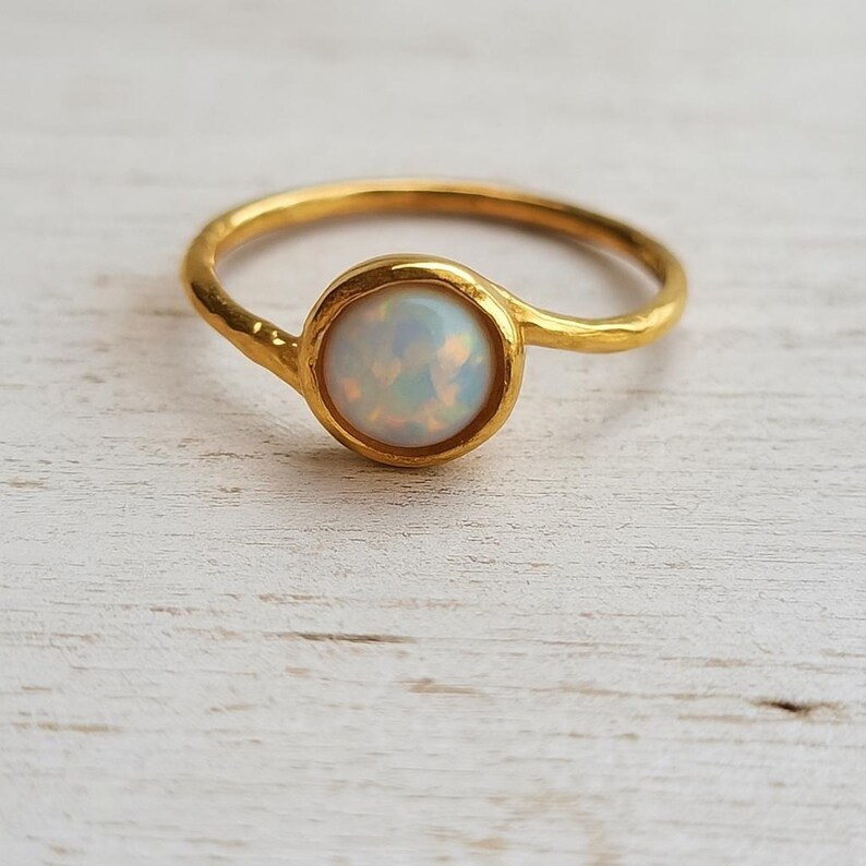 May include: A gold-colored ring featuring a round, iridescent opal stone. The opal is set in a bezel setting, and the band has a textured, hammered appearance. The ring is displayed on a light-colored wooden surface.