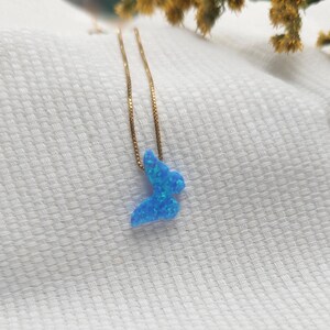 May include: A gold-colored chain necklace with a blue butterfly pendant. The butterfly pendant is a vibrant blue with a textured, opal-like appearance. The necklace is displayed on a white, textured surface.