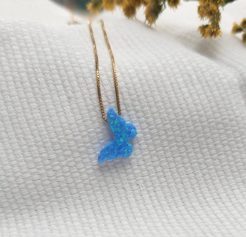 May include: A gold-colored chain necklace with a blue butterfly pendant. The butterfly pendant is a vibrant blue with a textured, opal-like appearance. The necklace is displayed on a white, textured surface.