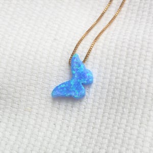 May include: A blue butterfly pendant necklace with a gold chain. The butterfly pendant has a marbled appearance with shades of light and dark blue. The necklace is displayed on a white textured surface.