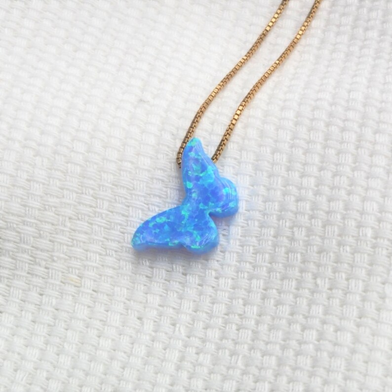 May include: A blue butterfly pendant necklace with a gold chain. The butterfly pendant has a marbled appearance with shades of light and dark blue. The necklace is displayed on a white textured surface.