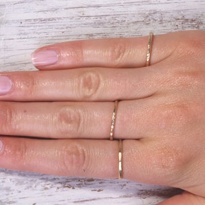 Pu&ograve; includere: Una mano con quattro anelli sottili color oro. Gli anelli presentano design strutturati e lisci. Le unghie della persona sono dipinte con uno smalto rosa chiaro. Lo sfondo &egrave; una superficie di legno bianco.