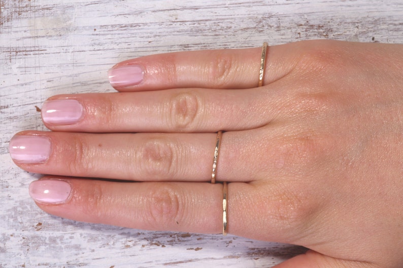 Pu&ograve; includere: Una mano con quattro anelli sottili color oro. Gli anelli presentano design strutturati e lisci. Le unghie della persona sono dipinte con uno smalto rosa chiaro. Lo sfondo &egrave; una superficie di legno bianco.