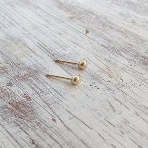 May include: Two gold ball stud earrings on a white wooden surface.