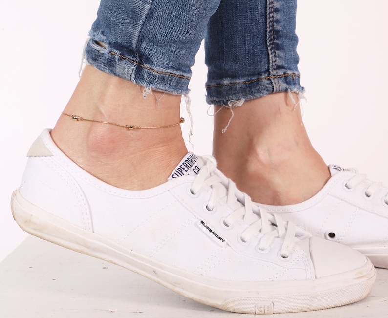 May include: A gold anklet with small round beads on a woman's ankle. The anklet is worn with white sneakers and blue jeans.