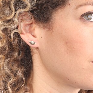May include: A close-up of a person's ear with a small gold leaf earring. The earring is a simple design with a textured surface.